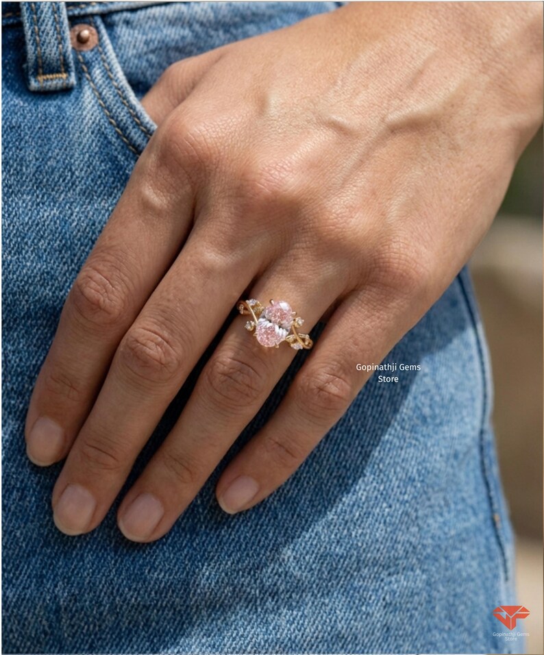 May include: A gold ring featuring two oval-cut pink gemstones. The ring has a delicate, ornate design with smaller accent stones. The ring is displayed on a hand, with a denim background.