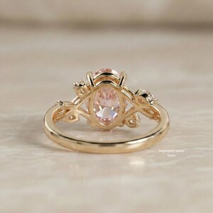May include: A gold ring featuring an oval, light pink gemstone. The ring has a delicate, floral design on the sides and around the gemstone. The ring is set on a light-colored surface.