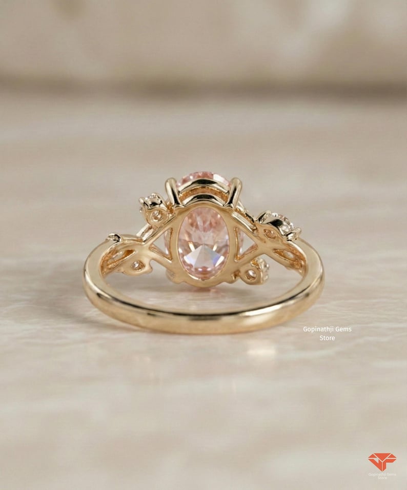 May include: A gold ring featuring an oval, light pink gemstone. The ring has a delicate, floral design on the sides and around the gemstone. The ring is set on a light-colored surface.