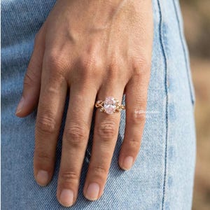 May include: A gold ring with an oval, light pink gemstone. The ring features a delicate, intertwined design with small accent stones. The ring is on a person's finger, set against a backdrop of blue denim.
