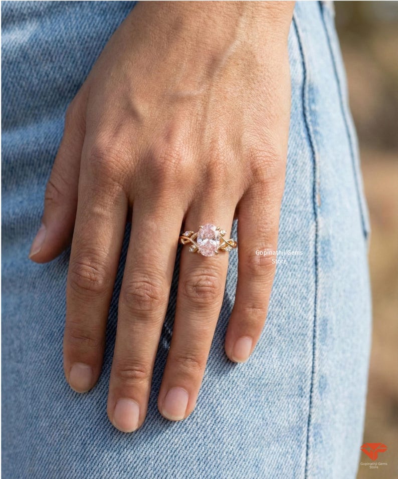 May include: A gold ring with an oval, light pink gemstone. The ring features a delicate, intertwined design with small accent stones. The ring is on a person's finger, set against a backdrop of blue denim.
