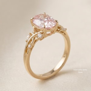 May include: A gold engagement ring featuring a large, oval-cut pink gemstone. The ring has a delicate, floral-inspired design with smaller accent stones. The band is smooth and polished, and the overall aesthetic is elegant and romantic.