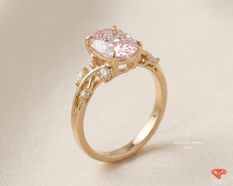 May include: A gold engagement ring featuring a large, oval-cut pink gemstone. The ring has a delicate, floral-inspired design with smaller accent stones. The band is smooth and polished, and the overall aesthetic is elegant and romantic.