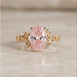 May include: A gold ring featuring an oval, light pink gemstone as the centerpiece. The ring has a floral design with small, clear accent stones. The ring is set on a light-colored surface. The text "Gopinathji Gems Store" is visible.