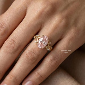 May include: A gold ring featuring a large, oval-cut pink gemstone as the centerpiece. The ring has a delicate, vine-like design with small accent stones. The ring is on a person's finger. The text "Gopinathji Gems Store" is visible.