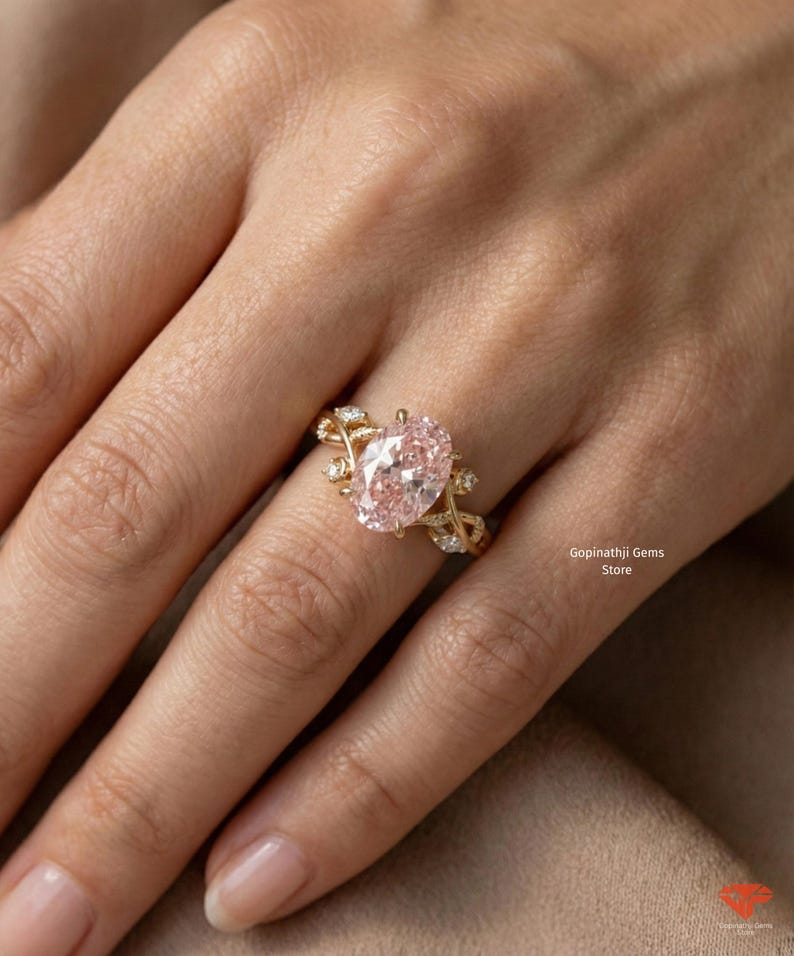 May include: A gold ring featuring a large, oval-cut pink gemstone as the centerpiece. The ring has a delicate, vine-like design with small accent stones. The ring is on a person's finger. The text "Gopinathji Gems Store" is visible.