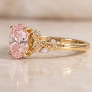 May include: A gold ring featuring a large, oval-cut pink gemstone. The ring has a floral design with small, clear accent stones. The band and floral accents are gold. The ring is displayed on a light-colored surface.