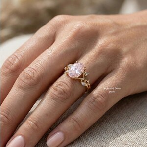 May include: A gold ring featuring an oval, light pink gemstone as the centerpiece. The ring has a delicate, leaf-like design with smaller, clear gemstones. The ring is on a person's finger.