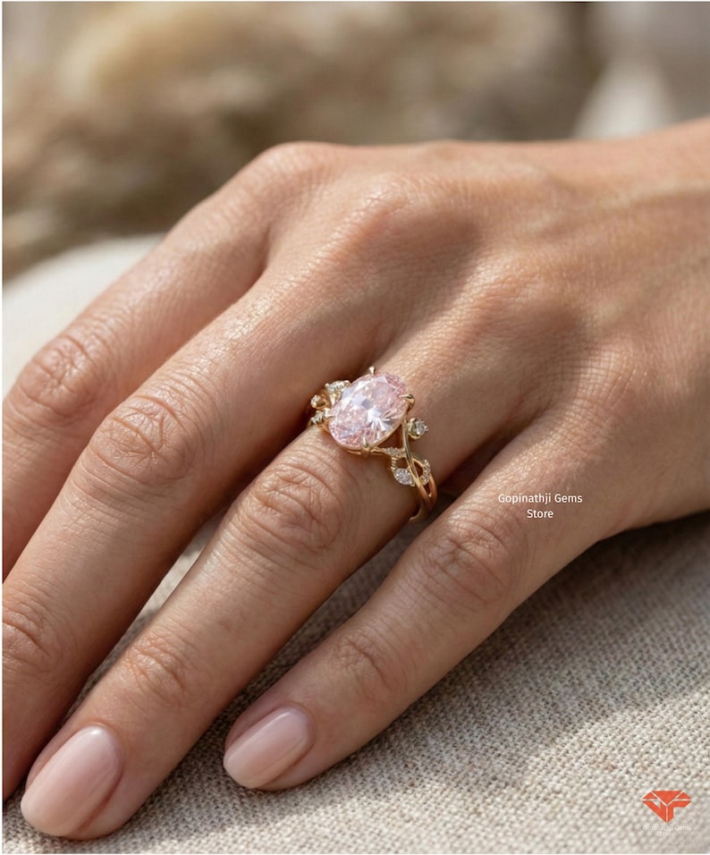 May include: A gold ring featuring an oval, light pink gemstone as the centerpiece. The ring has a delicate, leaf-like design with smaller, clear gemstones. The ring is on a person's finger.