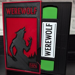 Op de afbeelding: Een zwarte en rode VHS-bandhoes met het woord "WEREWOLF" in vette letters. De hoes heeft een silhouet van een huilende wolf op een rode achtergrond. De VHS-band zelf is zwart met een groen venster en een wit label met de tekst "WEREWOLF".