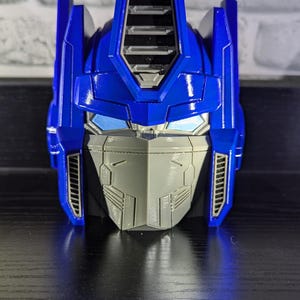 May include: A blue and gray 3D-printed model of Optimus Prime's head. The model features detailed design elements, including the faceplate, eyes, and helmet. The head is a vibrant blue with gray accents and is likely a collectible item.