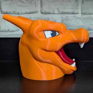 May include: An orange 3D-printed Charizard head, featuring a detailed design with white teeth, a red tongue, and blue eyes. The sculpture is set against a dark surface and a white brick wall background.