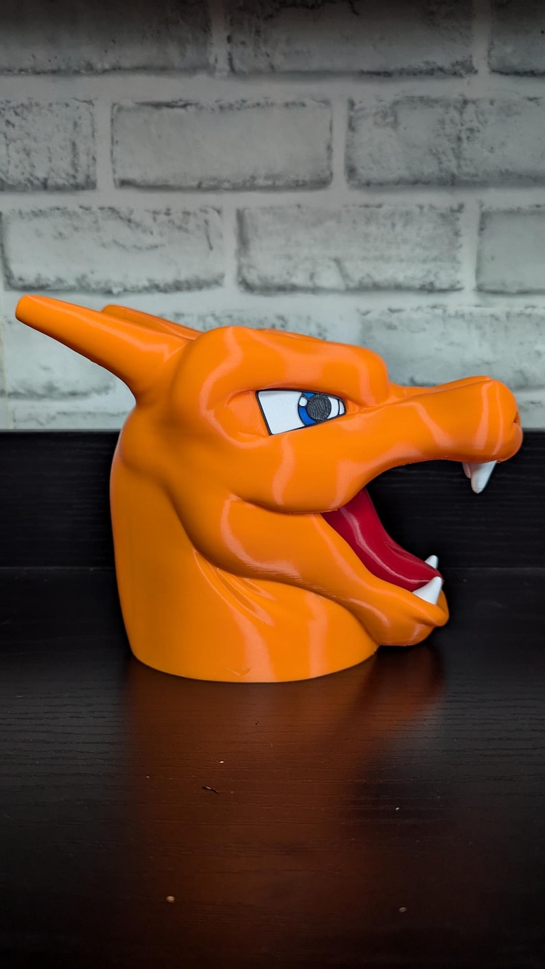 May include: An orange 3D-printed Charizard head, featuring a detailed design with white teeth, a red tongue, and blue eyes. The sculpture is set against a dark surface and a white brick wall background.
