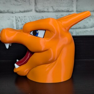 May include: An orange, 3D-printed Charizard head, featuring white fangs, a red tongue, and blue eyes. The stylized design is a popular character from the Pok&eacute;mon franchise. The head is a solid, glossy orange color.