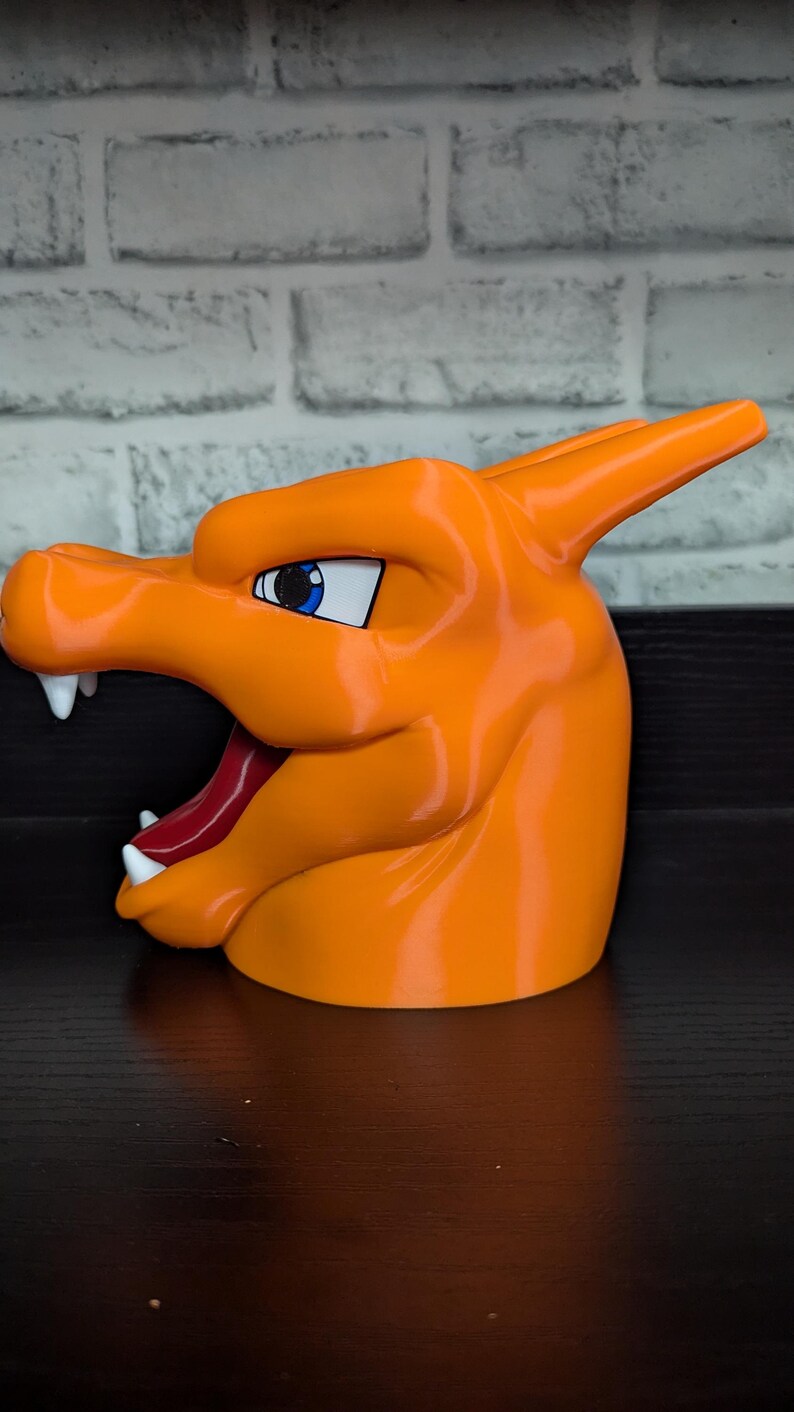 May include: An orange, 3D-printed Charizard head, featuring white fangs, a red tongue, and blue eyes. The stylized design is a popular character from the Pok&eacute;mon franchise. The head is a solid, glossy orange color.