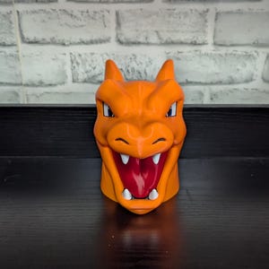 May include: Orange ceramic mug shaped like a Charizard's head, featuring a detailed face with white teeth, a red mouth, and blue eyes. The mug has two small horns and is set against a white brick wall.