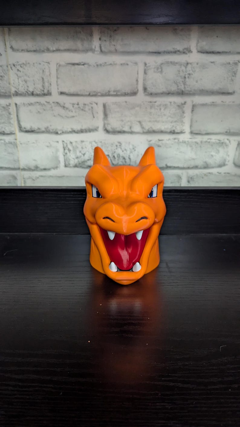 May include: Orange ceramic mug shaped like a Charizard's head, featuring a detailed face with white teeth, a red mouth, and blue eyes. The mug has two small horns and is set against a white brick wall.
