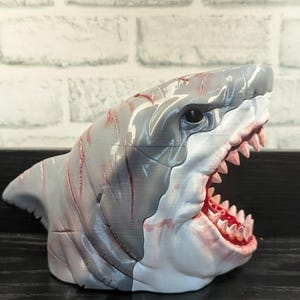 May include: A gray and white shark head sculpture with an open mouth revealing pink gums and sharp teeth. Red streaks are painted on the shark's head. The sculpture is displayed on a black surface against a white brick wall.