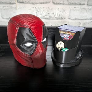 May include: A red and black Deadpool mask and a black card holder with dice. The mask is a detailed replica with white eye shapes. The card holder has a Deadpool image and holds cards and dice.
