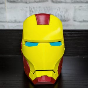 May include: A yellow and red Iron Man helmet replica with blue eye slits. The helmet is displayed on a dark surface against a white brick wall background. The helmet is a replica of the superhero's mask.
