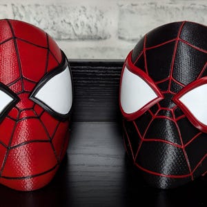 Puede incluir: Se muestran dos máscaras de superhéroe, una roja y otra negra. La máscara roja presenta un patrón de telaraña y formas de ojos blancos con contornos negros. La máscara negra tiene un diseño similar con líneas de telaraña rojas y formas de ojos blancos.