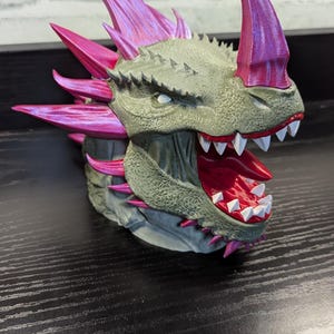 Puede incluir: Escultura detallada de una cabeza de dragón con la boca abierta, mostrando afilados dientes blancos y un interior rojo. El dragón presenta cuernos y púas rosas, con un exterior texturizado verde-gris.