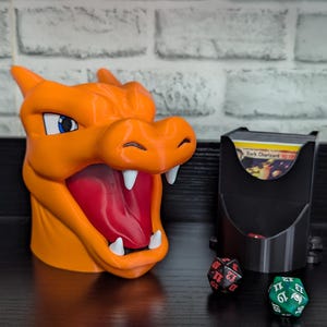 May include: An orange Charizard-themed card holder with a red interior and white teeth. A black card holder and three dice are also present. The card holder is designed to look like the Pokemon Charizard.