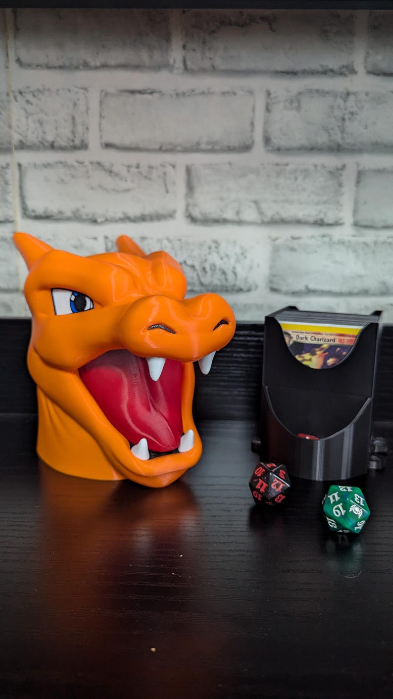 May include: An orange Charizard-themed card holder with a red interior and white teeth. A black card holder and three dice are also present. The card holder is designed to look like the Pokemon Charizard.