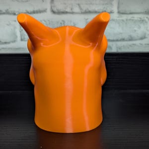 May include: Bright orange, 3D-printed sculpture of a horned creature. The sculpture has a smooth, glossy finish and a cylindrical base. The horns are cylindrical and point upwards. The sculpture is set against a black surface and a white brick wall.