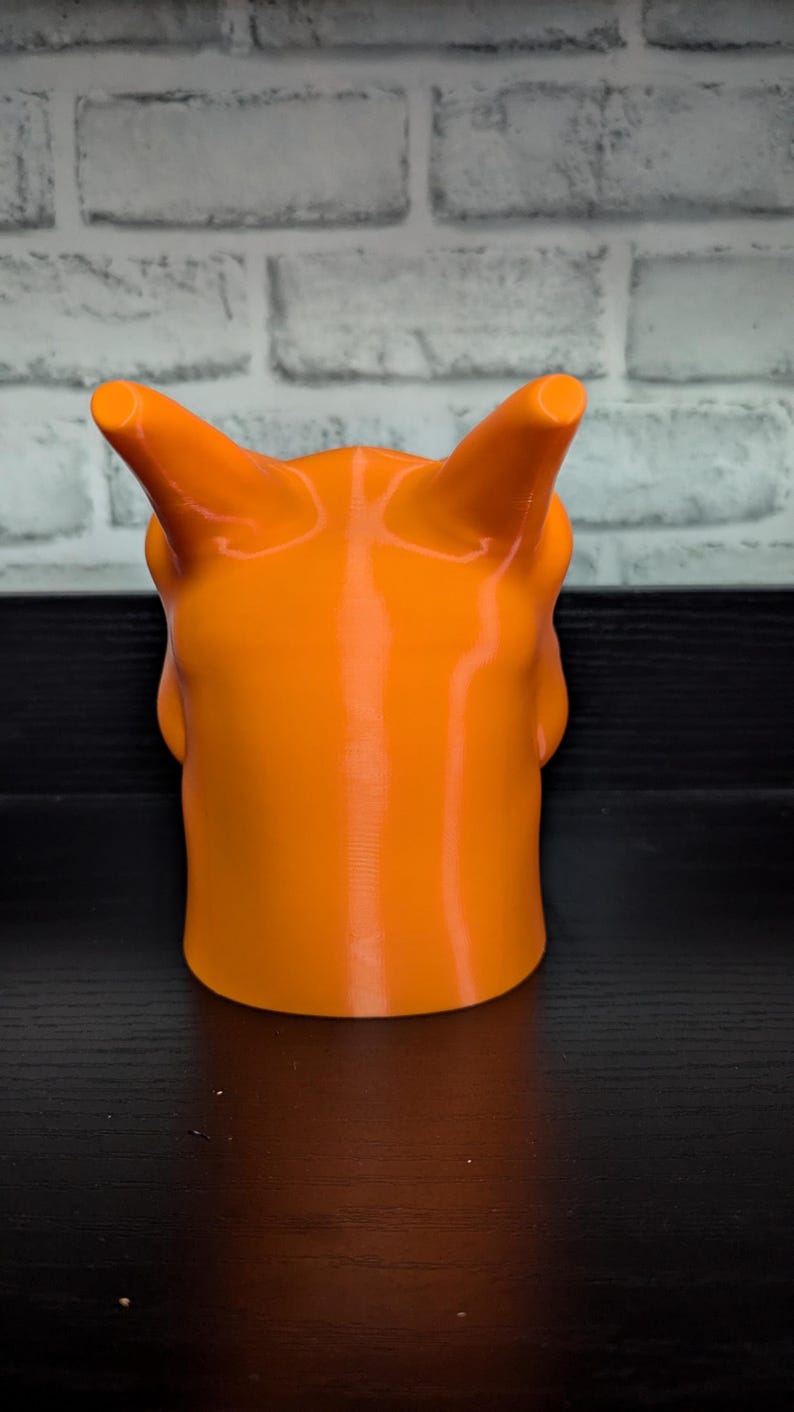 May include: Bright orange, 3D-printed sculpture of a horned creature. The sculpture has a smooth, glossy finish and a cylindrical base. The horns are cylindrical and point upwards. The sculpture is set against a black surface and a white brick wall.