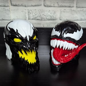 May include: Two detailed, sculpted masks, one with a yellow mouth and eyes, the other with a red tongue and sharp white teeth. Both masks are black and white, resembling comic book characters.