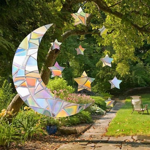 May include: A stained glass crescent moon and stars hanging from a tree branch in a garden setting. The moon and stars are iridescent and have a rainbow effect. There are two chairs in the background.