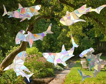 Puede incluir: Un conjunto de siete pegatinas de ventana de tiburón de vidrio tintado iridiscente. Las pegatinas tienen forma de tiburón y presentan un arcoíris de colores. Las pegatinas se muestran en un césped verde con un árbol en el fondo.