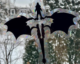 Puede incluir: Una silueta negra de un dragón con una persona montada en su espalda. El dragón está volando a través de un bosque nevado. La silueta está en una pegatina iridiscente.
