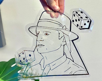 May include: A holographic sticker featuring a line drawing of a person wearing a hat and holding a dice. There is another dice in the background.