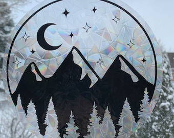 Può includere: Adesivo rotondo e olografico per finestre con una montagna nera, alberi, luna crescente e stelle. Il design è racchiuso in una cornice circolare. Lo sfondo è una scena invernale.