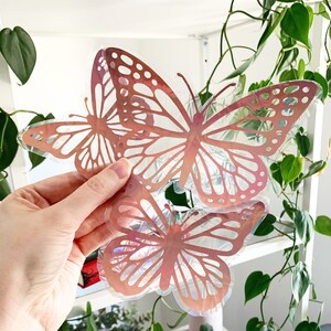 Könnte beinhalten: Zwei schimmernde rosa Schmetterlingsaufkleber mit holografischem Finish. Die Schmetterlinge sind übereinander gelegt und werden in einer Hand gehalten.
