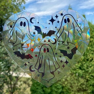 May include: An iridescent heart-shaped sticker featuring four ghosts, bats, stars, and crescent moons. The sticker is designed for a window or other smooth surface.