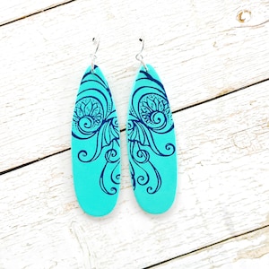 May include: A pair of turquoise teardrop earrings with a dark blue floral design. The earrings have a silver hook and are displayed on a white wooden surface. The earrings are a unique accessory.