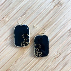 May include: A pair of rectangular black earrings with gold-colored trim and ear wires. Each earring features a gold swirling design on the lower half. The earrings are displayed on a light-colored wooden surface.