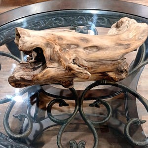 Op de afbeelding: Een groot stuk drijfhout met een lichtbruine kleur en een ruw, getextureerd oppervlak. Het drijfhout heeft een unieke vorm met verschillende gaten en rondingen. Het wordt tentoongesteld op een tafel met glazen blad en een decoratieve metalen voet.