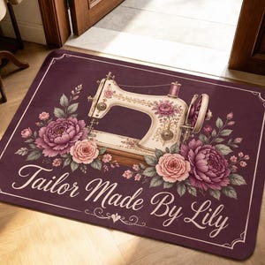 Op de afbeelding: Een rechthoekige donkerpaarse deurmat met een illustratie van een vintage naaimachine omringd door roze en paarse bloemstukken. De tekst "Tailor Made By Lily" staat in een wit cursief lettertype onderaan de mat gedrukt.