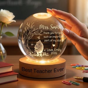 Op de afbeelding: Een heldere glazen bol met een ge&euml;tst Winnie de Poeh-ontwerp, verlicht door warm licht. De bol staat op een houten voet met de inscriptie "Best Teacher Ever". Tekst op de bol luidt "Mrs. Smith" en een bedankbericht, met "Love, Emily 2026".