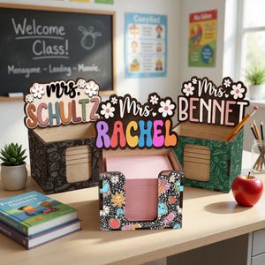 Könnte beinhalten: Holz-Schreibtisch-Organizer mit Lehrernamen, darunter "Mrs. Schultz", "Mrs. Rachel" und "Mrs. Bennet". Die Organizer haben ein Blumendesign und sind mit Mustern zum Thema Schule verziert. Ein roter Apfel und Bleistifte sind ebenfalls zu sehen.