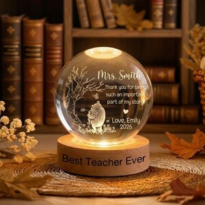 Op de afbeelding: Een heldere glazen bol met een ge&euml;tst ontwerp van een boom, een beer en de tekst "Mrs. Smith" en "Best Teacher Ever". De bol staat op een houten voet die van binnenuit verlicht is, waardoor een warme gloed ontstaat.