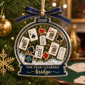 Könnte beinhalten: Ein transparenter, kugelförmiger Weihnachtsschmuck mit dunkelblauem Rand und einer dunkelblauen Schleife oben. Im Inneren befinden sich Miniatur-Spielkarten, ein Kompass-Plättchen und goldene Sterne. Text: 2026 und THE YEAR I LEARNED bridge.