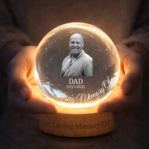 May include: A clear glass snow globe featuring a photograph of a man, inscribed with "DAD (1921-2022)". The globe rests on a wooden base, engraved with "In Loving Memory Of". The globe is held in cupped hands, with a warm glow emanating from within.