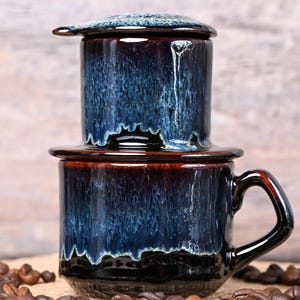 May include: A ceramic coffee filter and mug set with a dark blue and brown glaze. The coffee maker is stacked on top of the mug, with coffee beans scattered around the base. The set has a glossy finish.