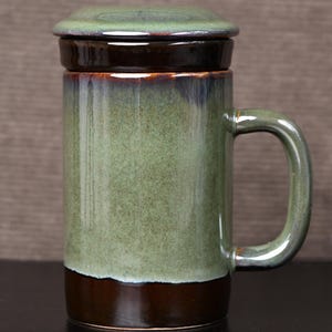 Puede incluir: Una taza de cerámica con tapa, con un esmalte verde y marrón. La taza tiene un asa y una forma cilíndrica. La tapa es plana y encaja perfectamente. La taza mide aproximadamente 15 cm de alto.