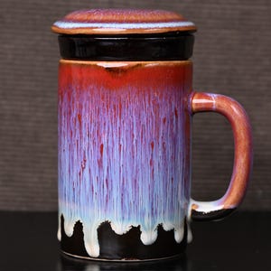 May include: A ceramic mug with a lid, featuring a vibrant color gradient. The mug transitions from red at the top to purple and blue, with white drips at the base. It has a brown handle and a black base.